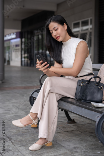 A gorgeous Asian female CEO is checking emails on her phone while sitting on a bench outdoors.