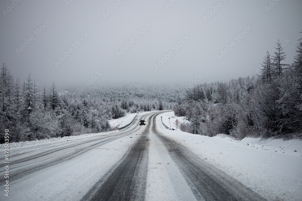 Fototapeta premium Photo of winter road during the snowfall in Magadan, Russia. Snow showers on trees and hills. Fog and haze, low visibility due to snowstorm. Extreme weather conditions. Car approaching in the distance