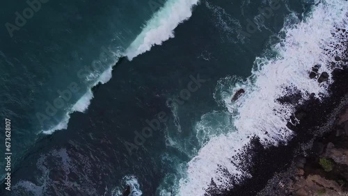 Beautiful cinematic top down aerial of deap clear blue ocean sea waves rolling to black rocky shore. Drone bird's eye view aerial, flying down photo