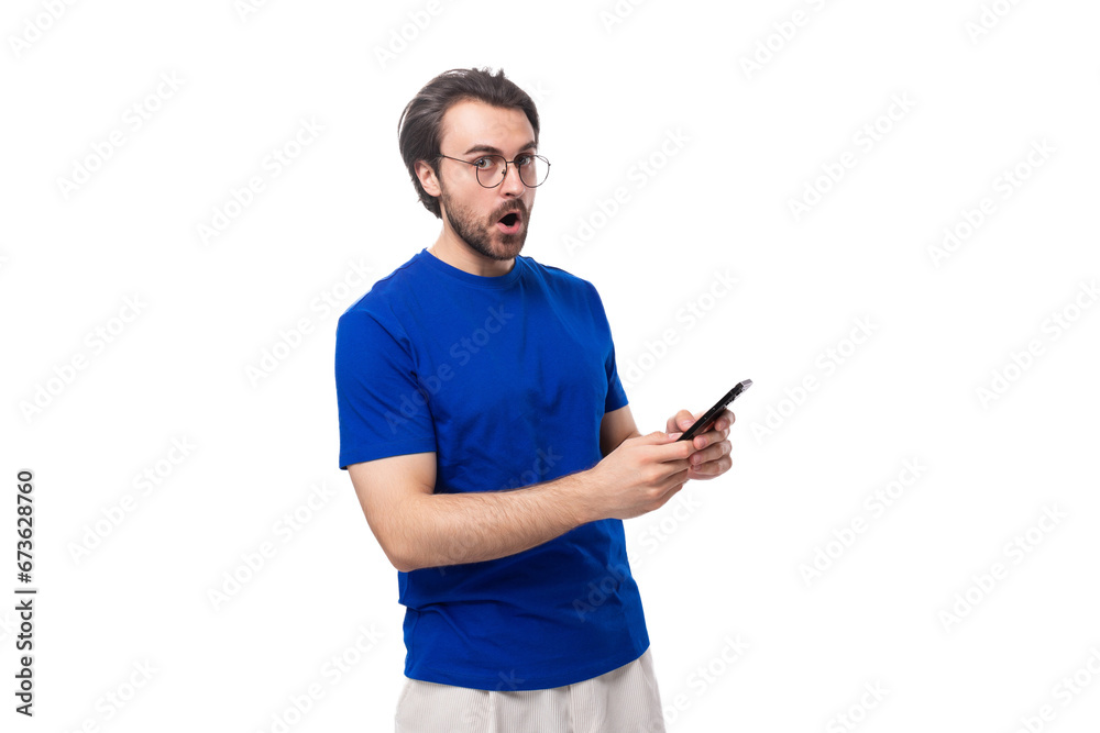 A 30-year-old European brutal guy with black hair and a beard in a blue T-shirt holds a phone in his hand in surprise A 30-year-old European brutal guy with black hair and a beard in a blue T-shirt holds a phone in his hand in surprise