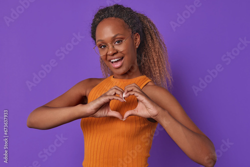 Young cute kind African American woman makes heart shape with hands and opens mouth and looks at screen inviting you on date or to go to cinema together stands posing on purple background.