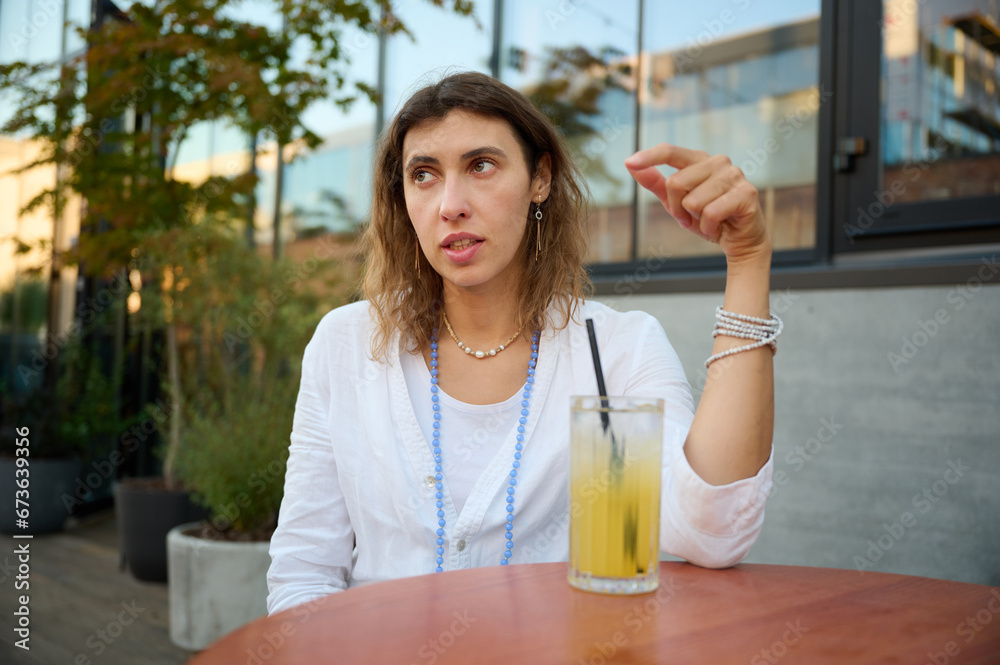 Young woman in a white shirt and a necklace around her neck on a bright sunny day in a cafe with greenery in the background is drinking fresh juice and actively talking about something Young woman in a white shirt and a necklace around her neck on a bright sunny day in a cafe with greenery in the background is drinking fresh juice and actively talking about something