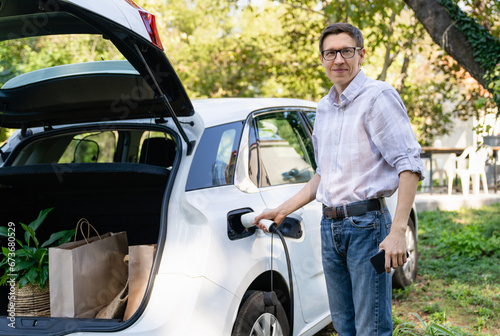 Man with shopping bag next to a charging electric car in the yard of a country house