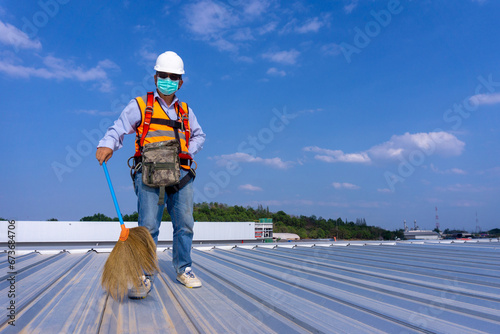 Worker wearing full safety body harness holding a broom in hand  working on roof top for cleaning metal roof sheet photo