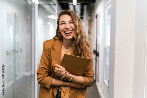 Happy businesswoman with clipboard standing in corridor at office photo
