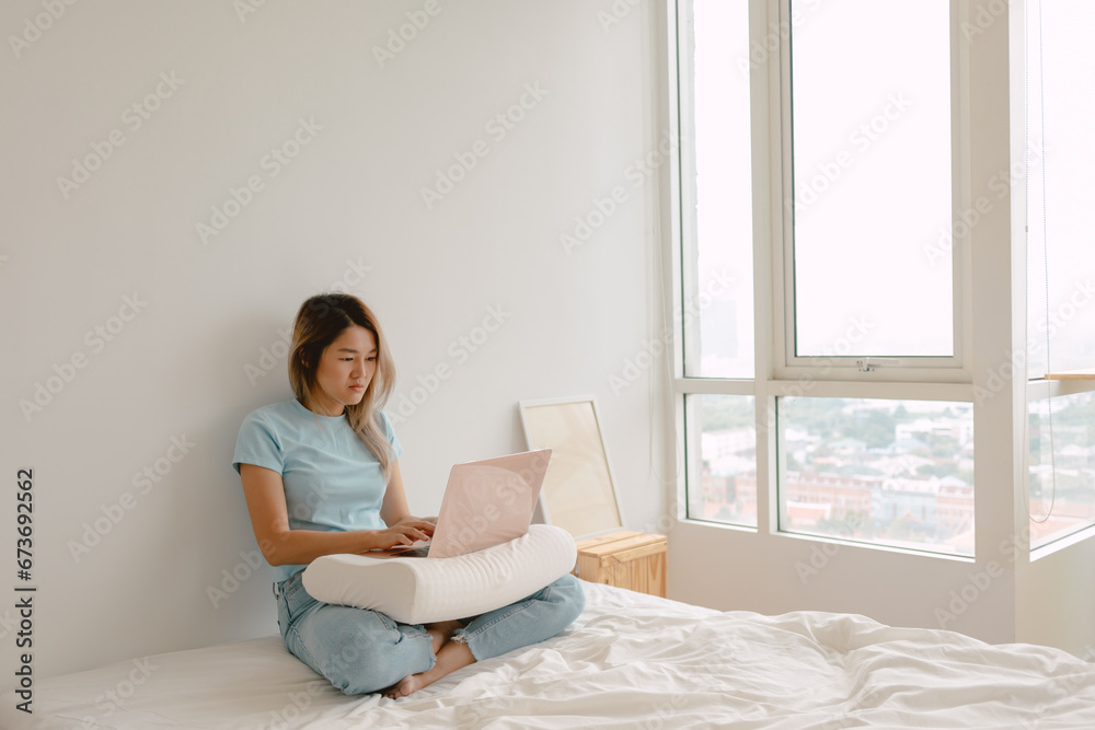 Asian Thai woman using laptop for working at home, putting computer on pillow, sitting on white bed, cozy chilling at apartment room alone.