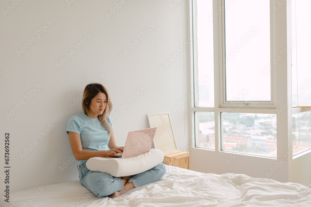 Asian Thai woman using laptop for working at home, putting computer on pillow, sitting on white bed, cozy chilling at apartment room alone. Asian Thai woman using laptop for working at home, putting computer on pillow, sitting on white bed, cozy chilling at apartment room alone.