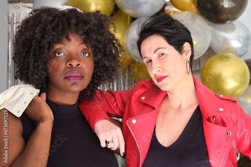Two multiracial serious women with leather jackets in a party with balloons.