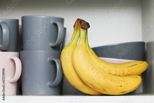 Bananas in kitchen cabinet. Coffee mugs. Tea cups. Milk soup bowls. Yellow fruits in kitchen. Open door kitchen cabinet with hidden bananas inside. Ripe bananas with small brown stains.