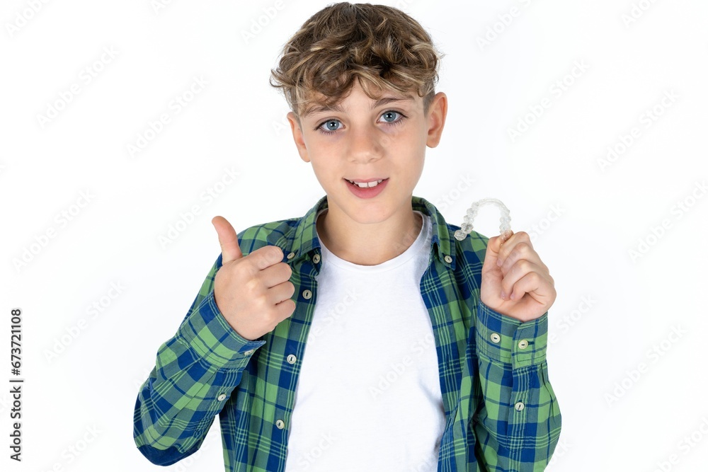 caucasian kid boy wearing plaid shirt holding an invisible braces aligner and rising thumb up, recommending this new treatment. Dental healthcare concept. caucasian kid boy wearing plaid shirt holding an invisible braces aligner and rising thumb up, recommending this new treatment. Dental healthcare concept.