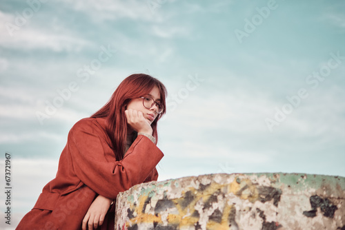 Pensive teenage girl in a red coat stands near a concrete podium with her hand propping her chin up.