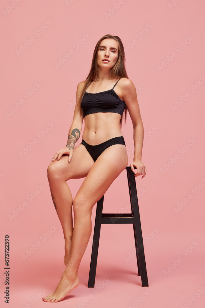 Yo9ugn attractive woman with fir, slim, muscular body sitting on chair in black underwear and posing against pink studio background. Concept of multi-ethnic beauty, spa, body acceptance, self-care