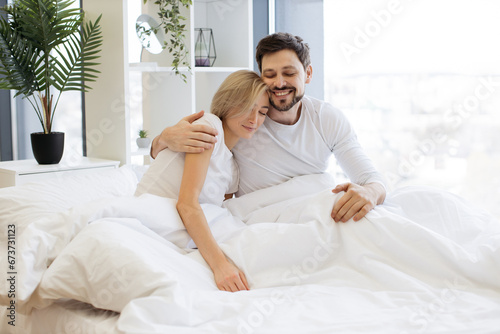 Loving caucasian couple in casual attire hugging after night sleep while still lying in bed of bright bedroom. Relaxed family man and woman starting new sunny day in modern flat.