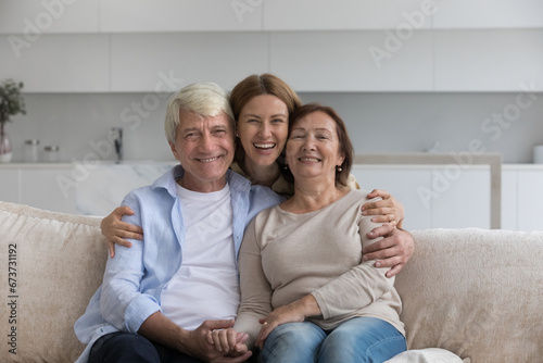 Happy beautiful adult daughter woman hugging elder mom and dad sitting on sofa together, embracing parents with love, tenderness, affection, looking at camera with toothy smile. Family portrait