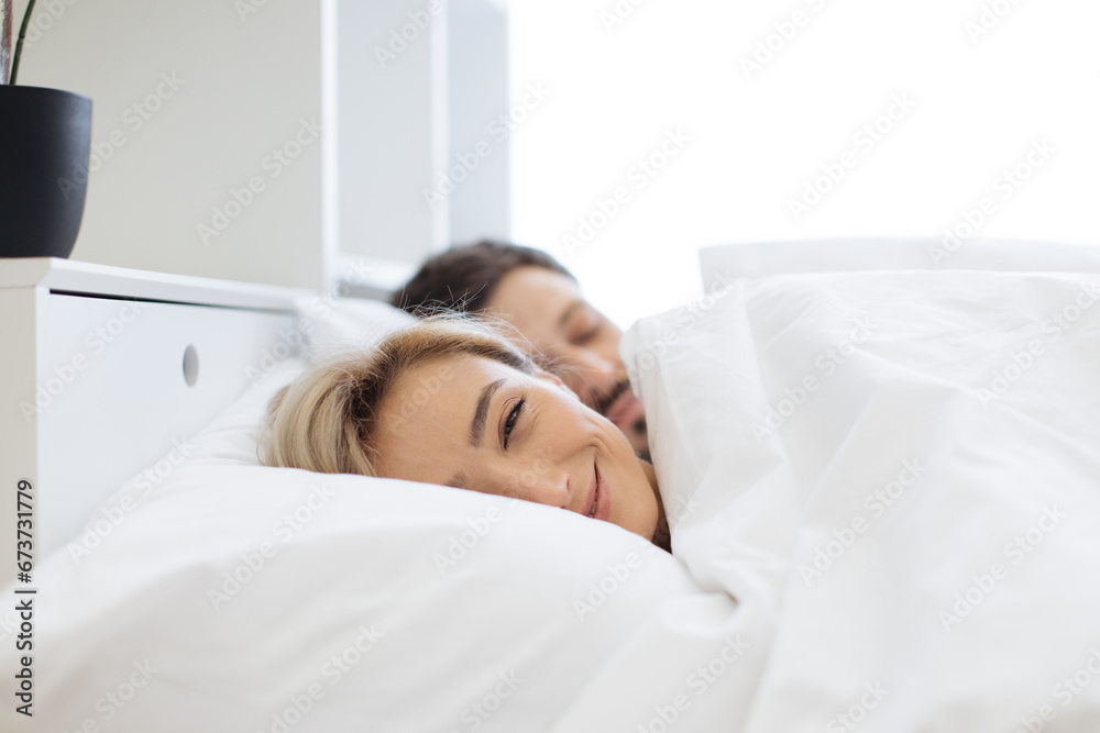 Pretty caucasian blond woman leaning on soft pillow already awake while her attractive husband still getting shuteye on bed in front of panoramic window. Happy couple spending morning together.