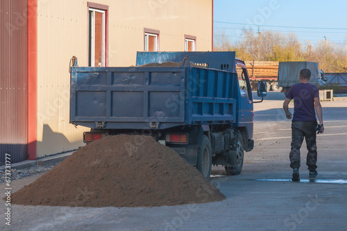 A small dump truck dumps sand from its back at a construction site. Yellow river sifted sand for preparing concrete mixture. A construction worker next to a car. Back view. Sunny day