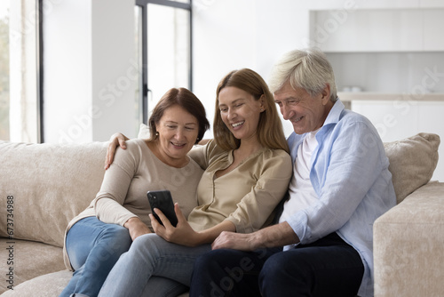 Cheerful elder couple of parents and adult child woman using mobile phone together, taking family selfie at home, sitting on sofa, posing, smiling, talking on video call on smartphone