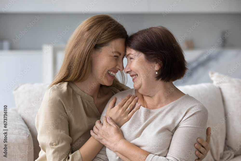 Positive beautiful adult daughter child hugging older mom, touching shoulders with love, gratitude, care, support, visiting parent at home on mothers day, looking at camera, smiling, laughing Positive beautiful adult daughter child hugging older mom, touching shoulders with love, gratitude, care, support, visiting parent at home on mothers day, looking at camera, smiling, laughing