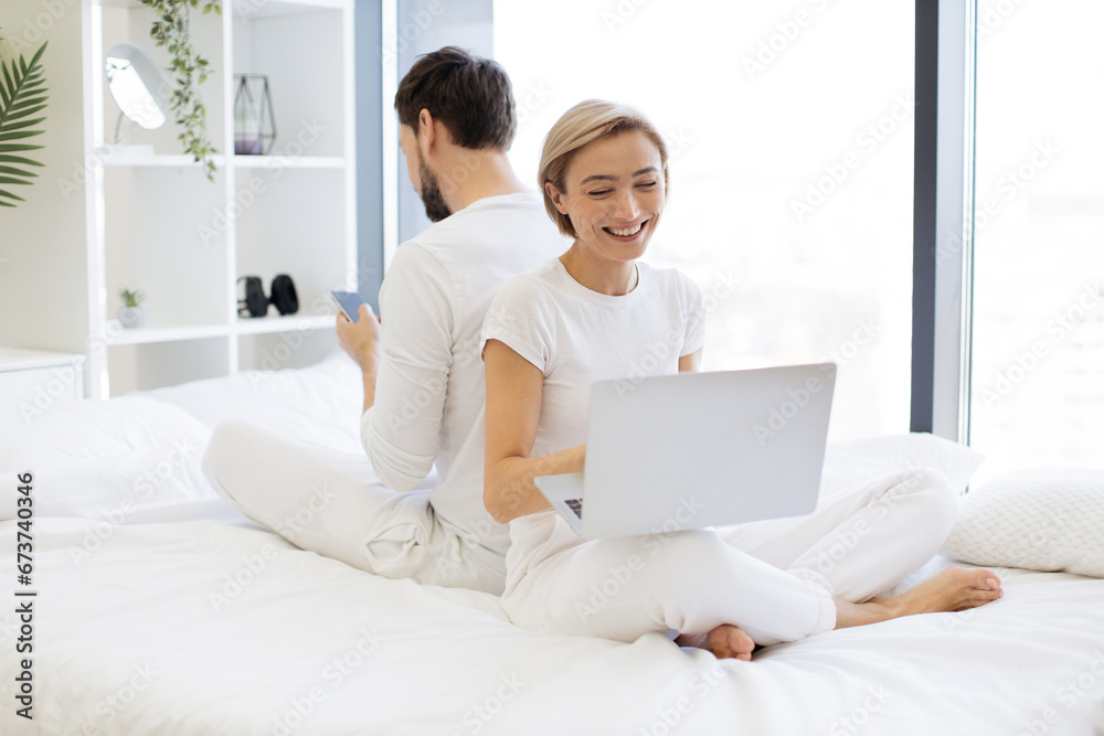 Smiling woman reading social updates using smartphone and serene man shopping online. Side view of caucasian couple in love working on modern gadgets while sitting back to back on bed. Smiling woman reading social updates using smartphone and serene man shopping online. Side view of caucasian couple in love working on modern gadgets while sitting back to back on bed.