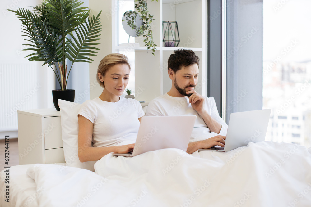 Side view of caucasian couple in love working on modern gadgets while lying under blanket at home. Smiling man reading social updates using laptop and serene woman shopping online. Side view of caucasian couple in love working on modern gadgets while lying under blanket at home. Smiling man reading social updates using laptop and serene woman shopping online.