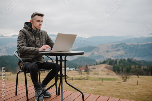 Man work on laptop sitting by table with great view mountains. Male working outdoors. Concept remote work or freelance lifestyle. Internet 5G. Workplace in country on backyard modern house in morning
