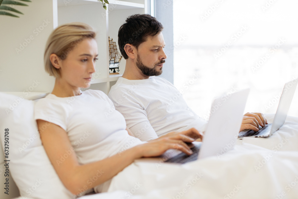 Attractive bearded caucasian man and pretty woman using digital devices while leaning against soft pillows in bed of studio room. Busy spouses doing their own business without disturbing each other.