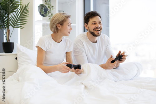 Side view of concentrated caucasian couple playing video games while lying under blanket at home. Smiling man and woman looking on tv have fun competing in video game using joysticks.