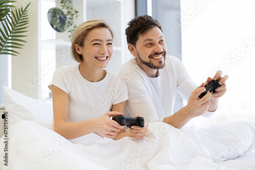 Cheerful amorous people looking at tv while spending time sitting on bed together playing video games in spacious apartment at weekend.