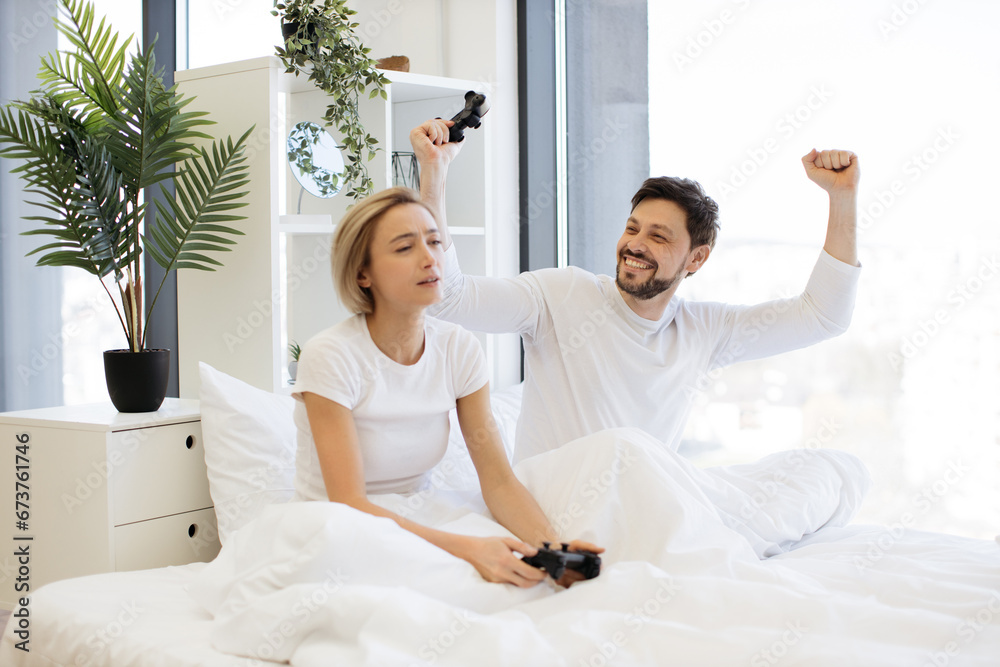 Sad woman holding joystick and looking at happy husband raised hands celebrating victory in video game. Side view of caucasian couple in love have fun competing in laptop game using joysticks on bed. Sad woman holding joystick and looking at happy husband raised hands celebrating victory in video game. Side view of caucasian couple in love have fun competing in laptop game using joysticks on bed.