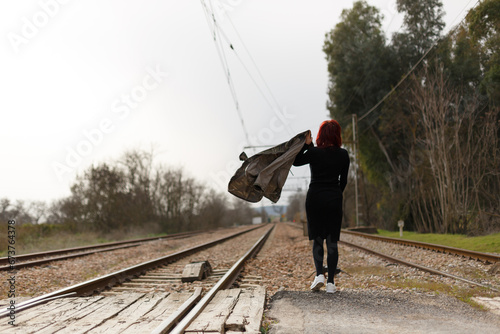 En las vías del tren una mujer caminando con chaqueta militar de espaldas, 