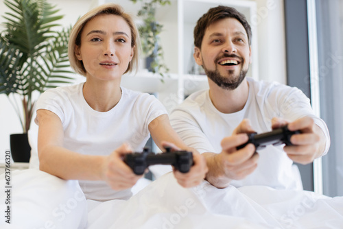 Close up of concentrated caucasian couple playing video games while lying under blanket at home. Smiling man and woman looking on tv have fun competing in video game using joysticks.