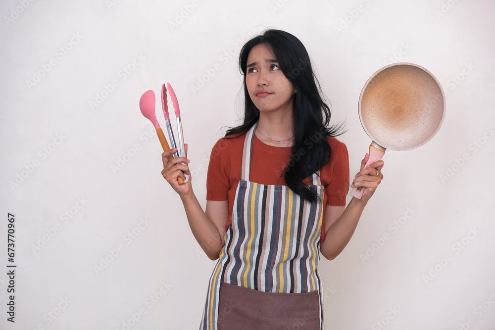Young housewife holding a frying pan and some kitchen utensils looking unsure what to cook Young housewife holding a frying pan and some kitchen utensils looking unsure what to cook