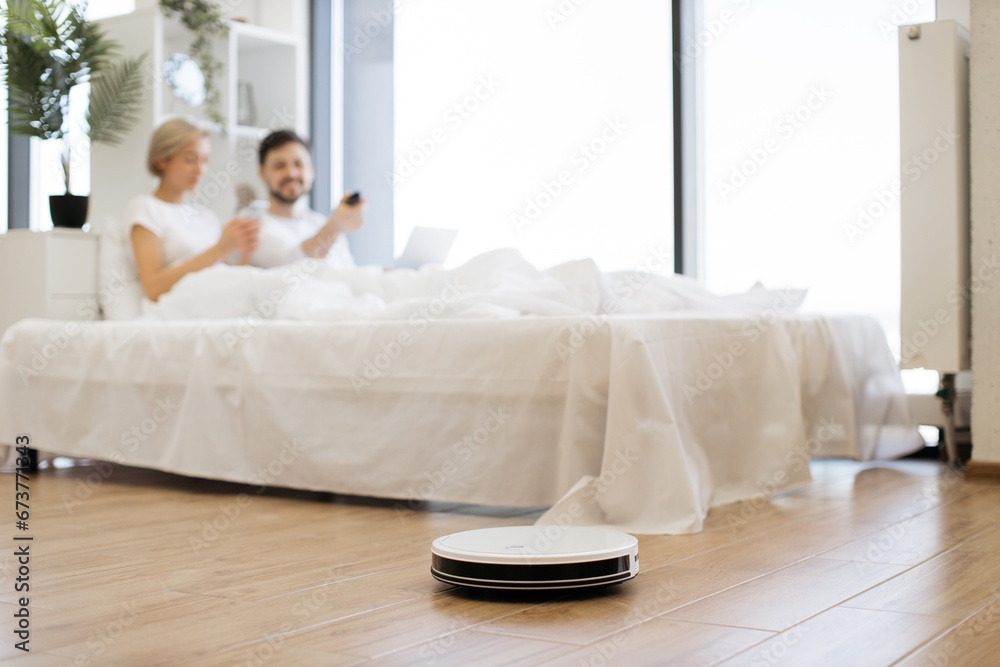 Selective view of modern robot vacuum cleaning wooden floor in bedroom while young multiracial couple watching video on laptop. Young family saving time for leisure by using electrical device. Selective view of modern robot vacuum cleaning wooden floor in bedroom while young multiracial couple watching video on laptop. Young family saving time for leisure by using electrical device.