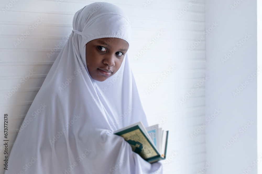 African Muslim child girl wears hijab abaya reading the Quran, leaning against white wall African Muslim child girl wears hijab abaya reading the Quran, leaning against white wall