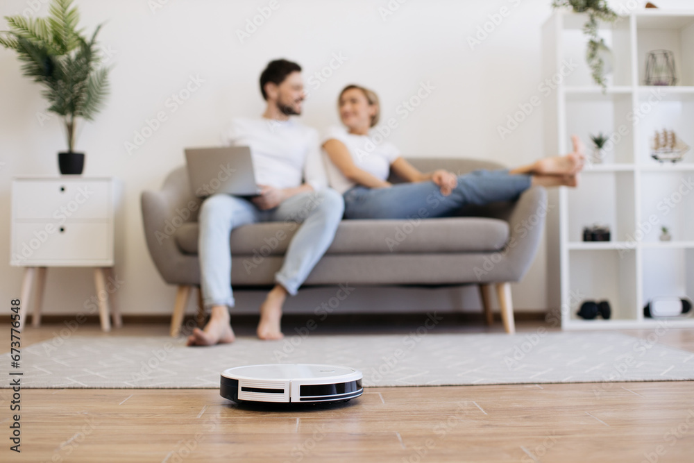 Automatic robot vacuum cleaning wooden floor while happy young couple resting on background. Two caucasians in love spending time for watching movie instead of households.