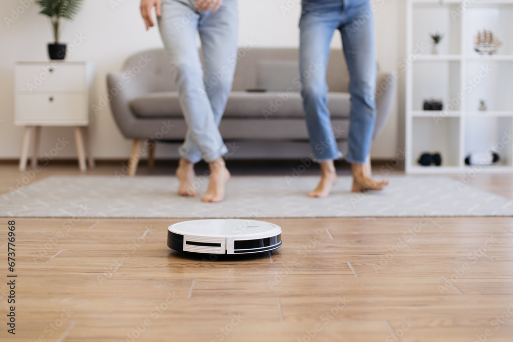 Close up view of modern cleaning robot vacuuming bare laminate floor with cropped view of male and female in background. Automatic gadget dusting off surface while wife and husband dancing for music.
