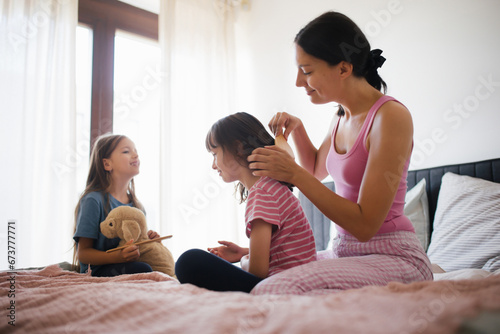 Mother with two daughters on a bed in the bedroom. Mother combs daughter's long hair with comb.Concept of Mother's Day and maternal love.