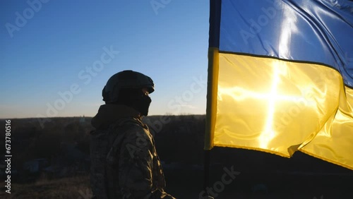 Ukrainian army girl stands with lifted national banner against background of sunset. Young female soldier in military uniform holds a waving flag of Ukraine to peak of hill. Victory of war. Dolly shot photo