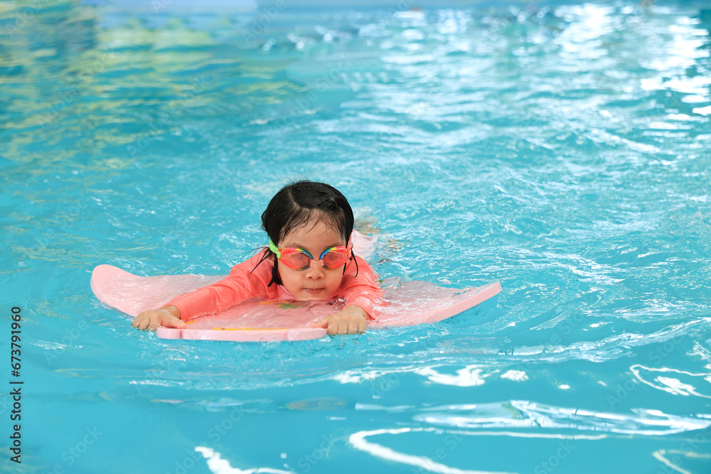 Smiling little girl learning to swim in pool with flutterboard during swimming class