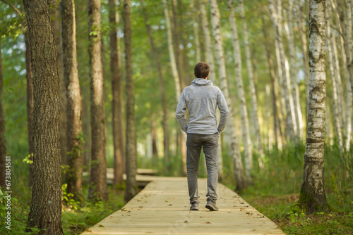 Young adult man walking on wooden trail at birch tree forest in beautiful autumn day. Spending time alone and enjoying freedom at nature. Back view. Peaceful atmosphere in nature.