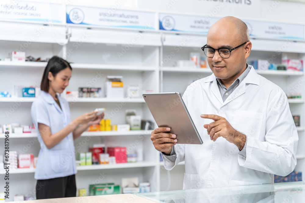 Happy handsome asian male pharmacist wearing eyeglasses and lab coat standing with tablet with coworker. He feels good, trustworthy and proud of his work in the pharmacy drugstore.