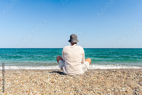 A man sits on the beach in the lotus position and looks at the sea. Meditating man in summer clothes on the beach.