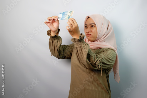 Unhappy beautiful Asian Muslim woman holding paper money isolated on white background. ​ photo