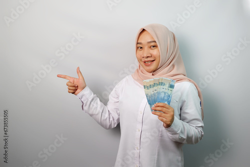 Smiling young Asian hijab woman holding paper money and pointing at banner advertisement standing over white background.