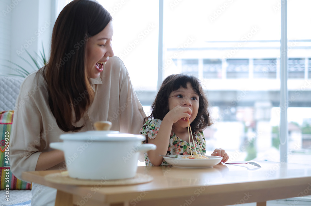 Young daughter picks up some spaghetti with her hands and eats it as her mother laughs happily. Young daughter picks up some spaghetti with her hands and eats it as her mother laughs happily.