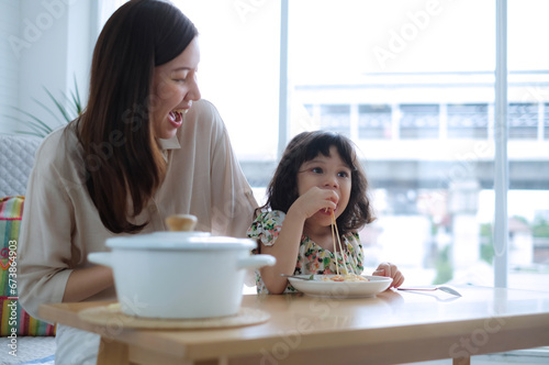 Young daughter picks up some spaghetti with her hands and eats it as her mother laughs happily.