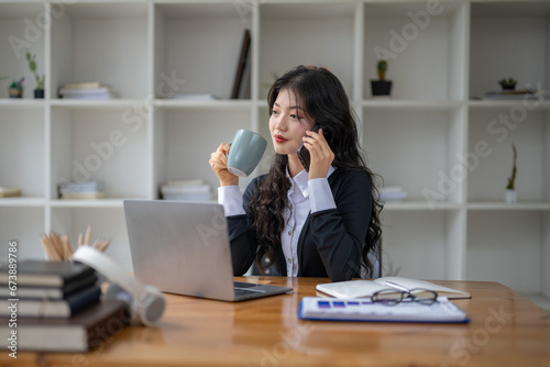 Freelance Asian woman talking on the phone and sipping coffee while working at the desk.