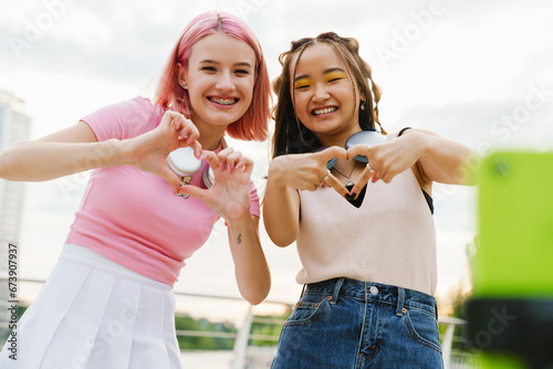 Two smiling girls recording videos with smartphone and showing heart gesture while standing on city waterfront