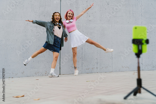 Two cheerful girls making dancing video with smartphone outdoors near gray wall photo