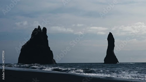 Huge sea stacks of the famous Black Beach in Iceland. Reynisfjara Black Sand Beach. Waves crash on the rocks and seagulls fly around the majestic rocks. Still shot in slow motion, 4k.